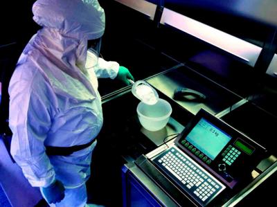 Fotoet viser en kvindelig laborant i et mørklagt laboratorium. Laboranten vejer et hvidt pulver af ned i en hvid plastikskål på en elektronisk vægt, der viser 0,3 kg. Laboranten er i hvid beskyttelsesdragt med hætte, visir og handsker.  