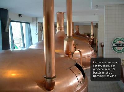 Fotoet viser fire store brygkedler i kobber i en bryggerihal med hvide fliser på vægge og gulv. På væggen til højre for brygkedlerne hænger et skilt for ølmærket Heineken.