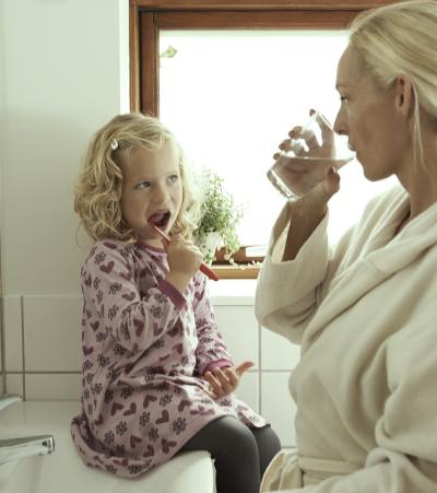 Foto viser en mor i morgenkåbe med sin lille lyshårede datter i lyserød blomstret kjole i badeværelset. Pigen sidder på håndvasken og børster tænder, mens hun ser på sin mor, der drikker af et glas vand. Imens har de lukket for vandet for at undgå vandspild.