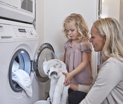 Fotoet viser en mor i beige cardigan, der sidder på knæ foran en vaskemaskine. Ved siden af står hendes lille lyshårede datter i lyserød blomstret kjole. De er i færd med at tage vasketøj ud af en fyldt vaskemaskine og lægge det ned i en hvid vasketøjskurv. Oven på vaskemaskinen står der en tørretumbler.