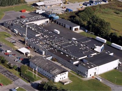 Billedet viser  et luftfoto af et stort hvidt fabriksanlæg med grå tage. Fabrikken ligger i landlige omgivelser med grønne arealer omkring. 
