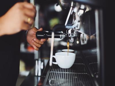 Fotoet viser en persons hænder, der er ved at brygge espresso kaffe fra et espressoanlæg i en hvid kop med hank. 
