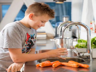 Fotoet viser en dreng i en grå T-shirt med og mørkt kort pjusket hår på ca. 12 år. Han er i færd med at tappe koldt vand fra køkkenhanen i et glas. På køkkenbordet ligger nogle gulerødder i forgrunden og bag ved vasken står en skål med grøn salat.