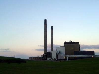 Fotoet viser et landskab med et kraftværk ved skumringstid. Kraftværket har to høje slanke skorstene, der rager højt op i luften. Ved siden af ligger de store kraftsværksbygninger. 