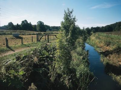 Foto viser et naturlandskab. Til venstre i billedet ses en indhegnet grøn græseng med en kildeplads og en cementring opført midt på. Det er en boring til indvinding af vand. Til højre i billedet ses et åløb med græsskråninger og småbuske langs åen