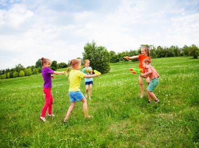 Fotoet viser en grøn græseng med mindre buske i baggrunden. Det er sommer og på engen leger fire piger og en dreng på en 10 – 12 år med store og små vandpistoler. De skyder med vand på hinanden, griner og har det sjovt. 